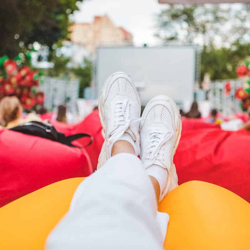 A relaxed view from the perspective of someone lounging on a bright yellow beanbag at an outdoor film screening. The person’s legs, dressed in white pants and white sneakers, are stretched out in front of them. In the background, people sit on red beanbags facing a large movie screen framed by trees and colorful decorations, creating a festive and comfortable atmosphere in a park or public space.