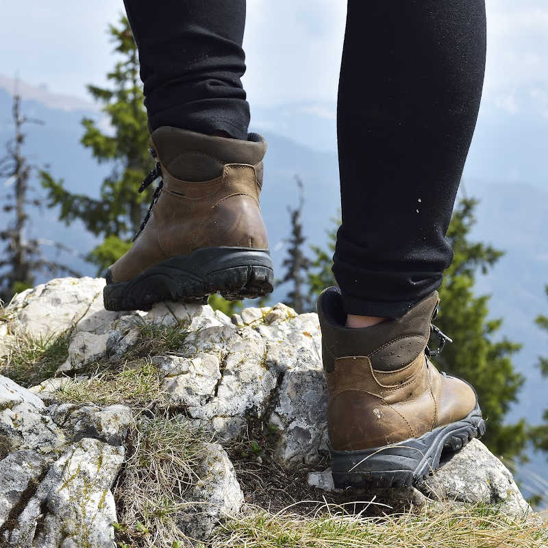 A close-up of a hiker's brown boots standing on rocky terrain with mountains and evergreen trees in the background, capturing a moment of outdoor exploration and adventure.