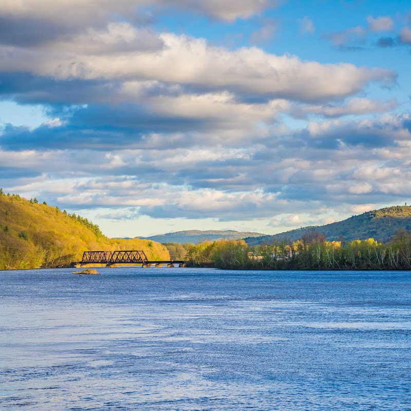 View of the Connecticut River with a truss bridge in the distance, surrounded by forested hills under a partly cloudy sky.