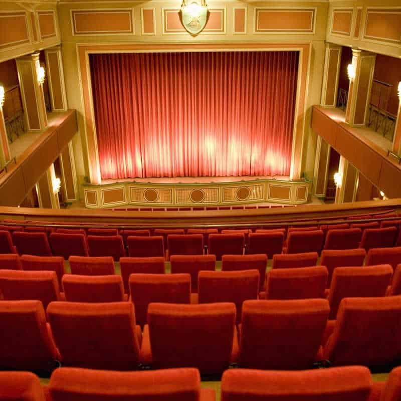 An elegant theater interior features rows of plush red seats facing a large stage framed by ornate paneling and glowing sconces. The stage curtain is drawn, casting a warm, golden light that enhances the rich tones of the room. Decorative balconies and classical architectural details contribute to the refined and inviting atmosphere.