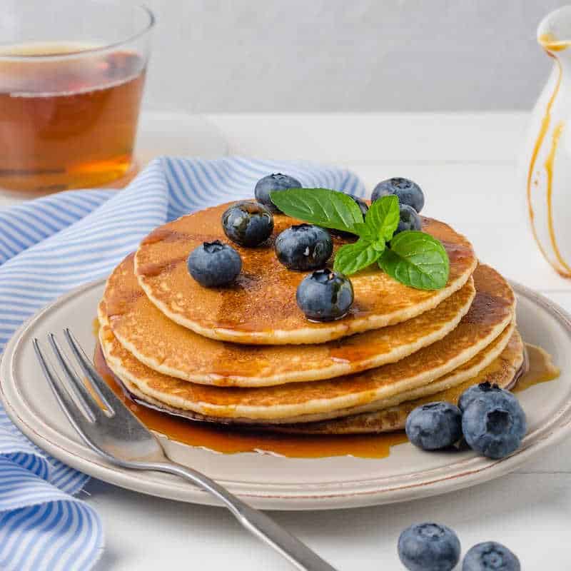 Stack of golden pancakes topped with fresh blueberries, mint leaves, and drizzled maple syrup on a white plate, with a fork beside it and a glass of tea in the background.