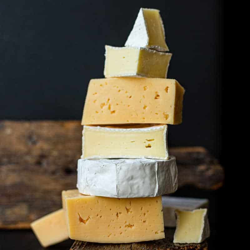 A stack of assorted cheese wedges, including soft and semi-soft varieties with white rinds and yellow interiors, arranged on rustic wooden boards against a dark background.