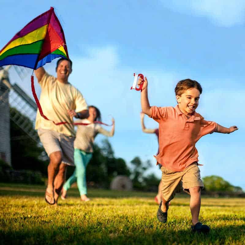 A young boy runs joyfully across the grass flying a rainbow kite, followed by smiling adults and another child.