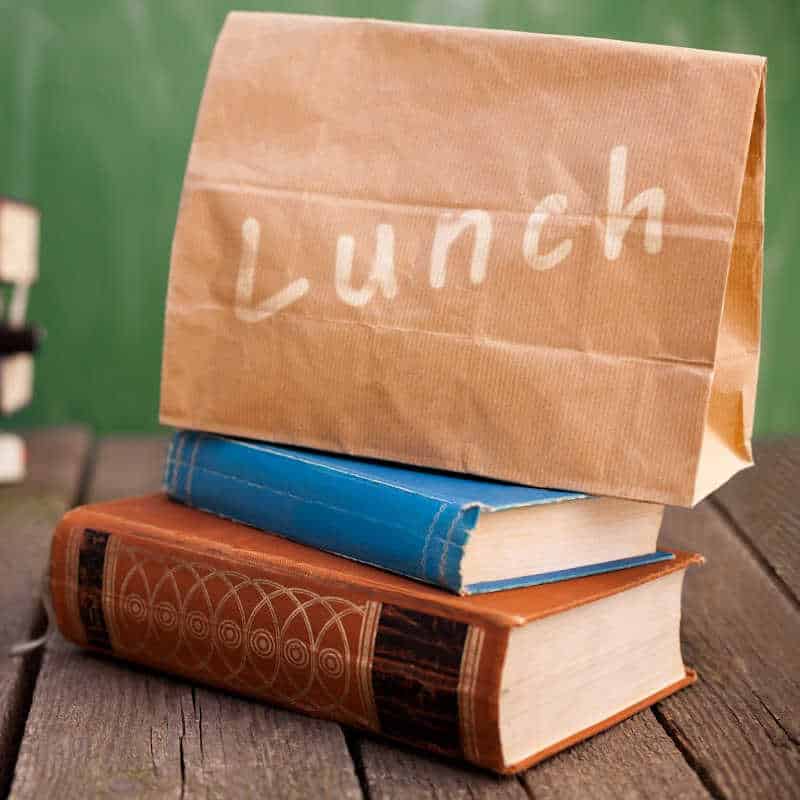 A brown paper lunch bag labeled 'Lunch' resting on top of two stacked hardcover books on a wooden table.