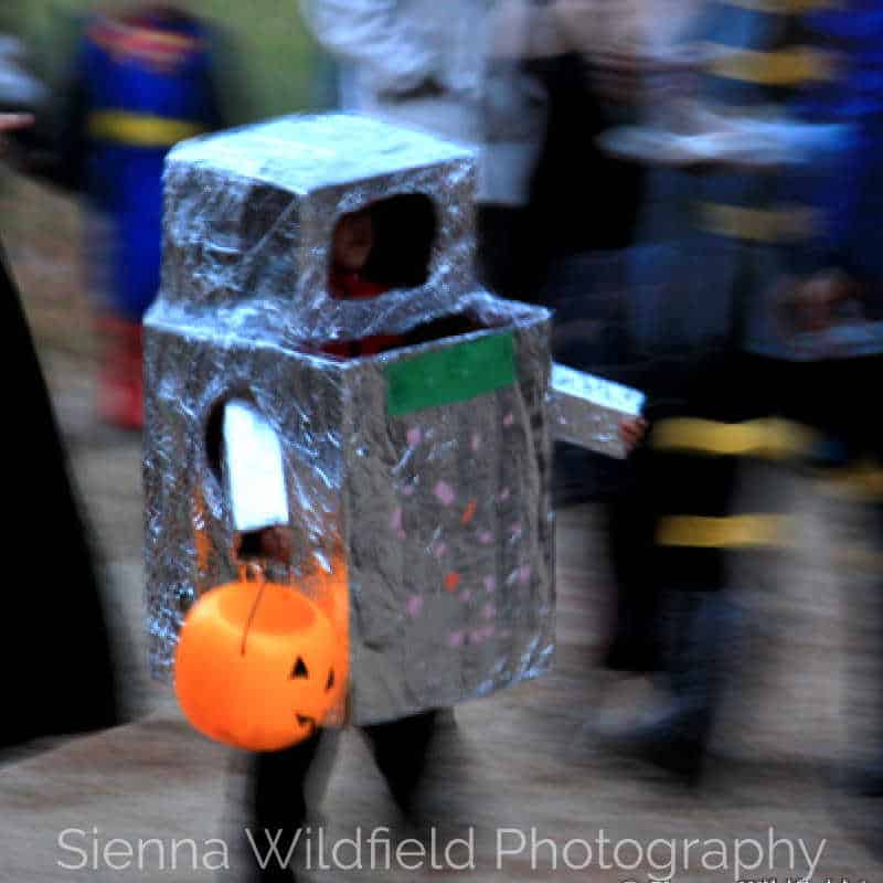 Child dressed in a homemade robot costume covered in foil, carrying a pumpkin bucket while trick-or-treating. (Photo credit: Sienna Wildfield)
