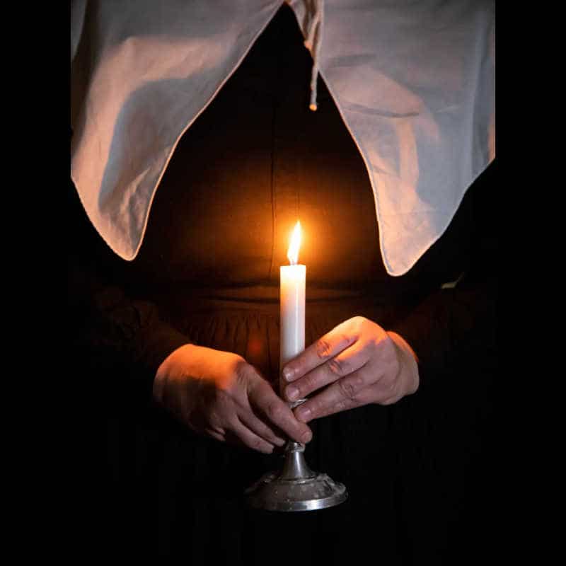 Person in period clothing holding a lit candle in the dark during an Old Sturbridge Village program.