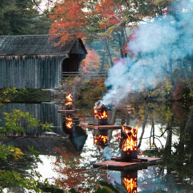 Row of floating bonfires reflecting on the water with a covered bridge and autumn trees in the background at Old Sturbridge Village.