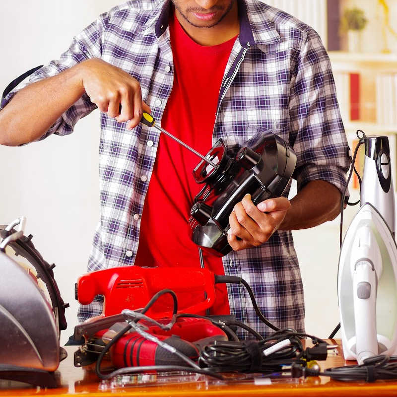 A person repairing a small household appliance with a screwdriver, surrounded by various tools and electronics on a table at a repair café.