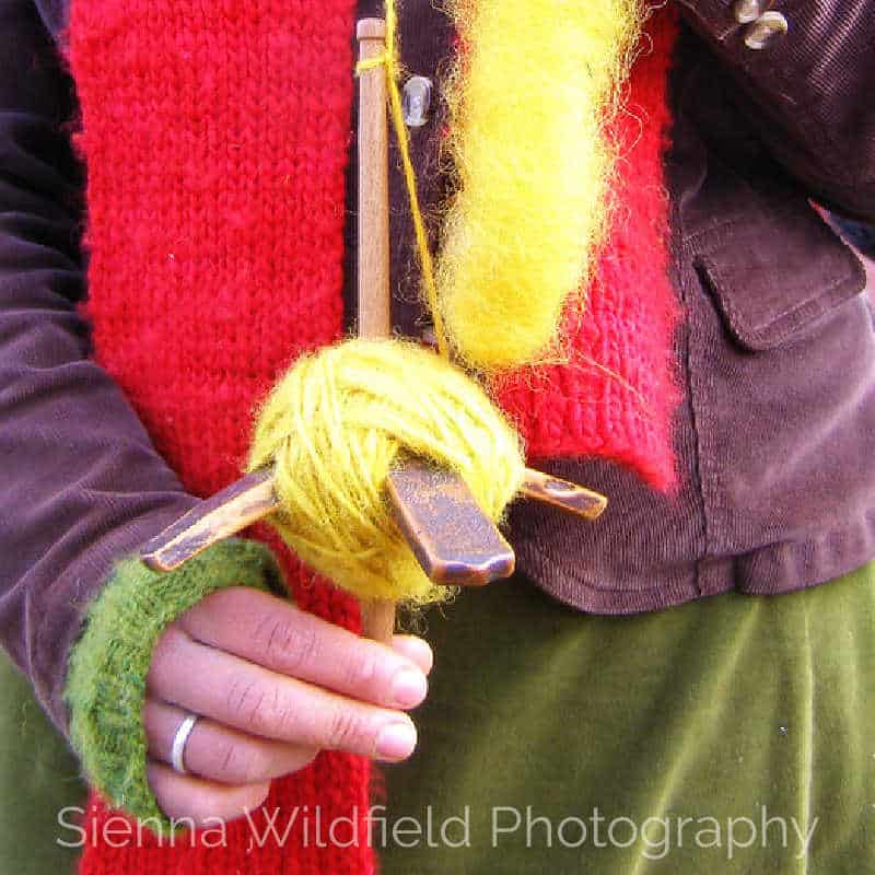 Person holding a wooden drop spindle with bright yellow yarn, wearing a red scarf and green mitten.