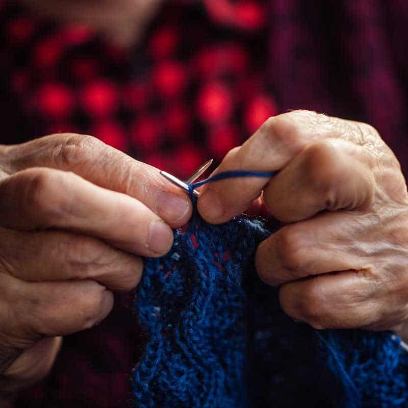 Close-up of elderly hands knitting with blue yarn, symbolizing craft traditions and intergenerational learning.