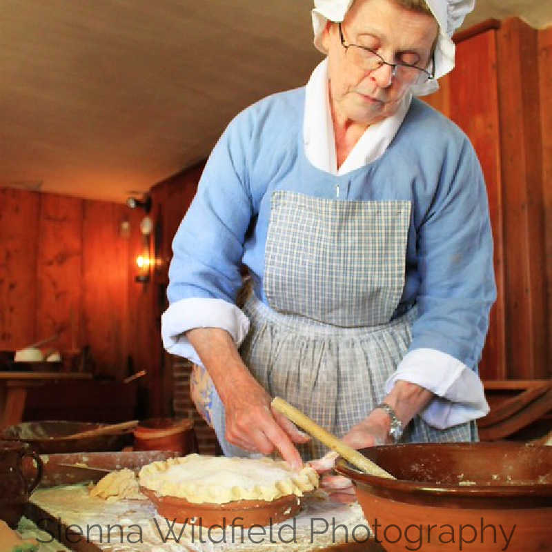 A woman in 18th-century clothing shapes dough in a colonial kitchen, wearing a cap and apron as she prepares food by hand.
