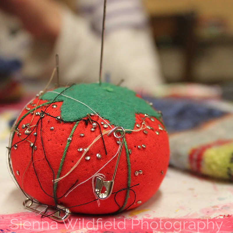 Close-up of a handmade tomato-shaped pincushion with sewing needles and thread on a worktable.