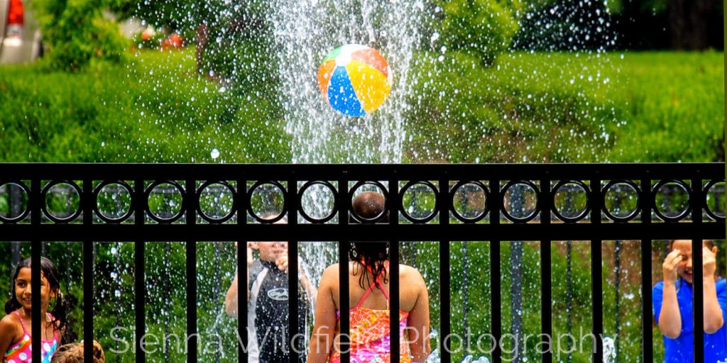 Beat the Heat in Western Massachusetts: Children play behind a black metal fence as water sprays around a colorful beach ball at the splash pad at Look Memorial Park in Florence, MA. (Photo credit: Sienna Wildfield)
