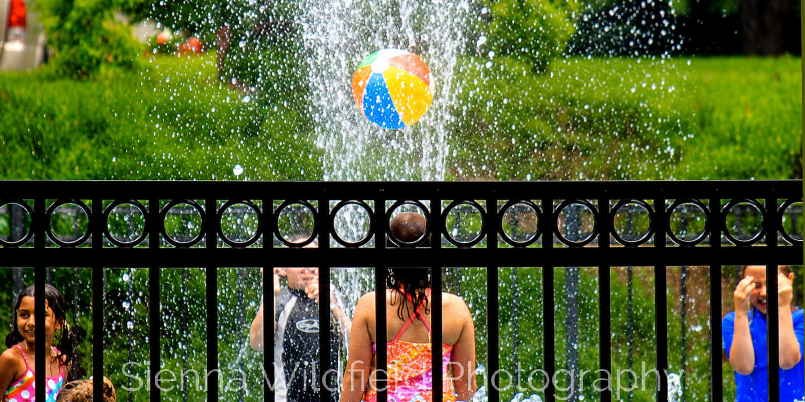 Beat the Heat in Western Massachusetts: Children play behind a black metal fence as water sprays around a colorful beach ball at the splash pad at Look Memorial Park in Florence, MA. (Photo credit: Sienna Wildfield)